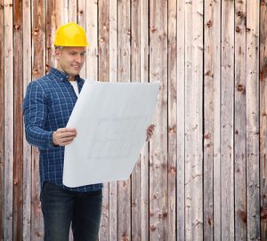 Photo of smiling male builder or manual worker in helmet with blueprint over wooden fence background