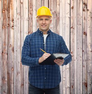 Photo of smiling male builder or manual worker in helmet with clipboard taking notes over wooden fence background